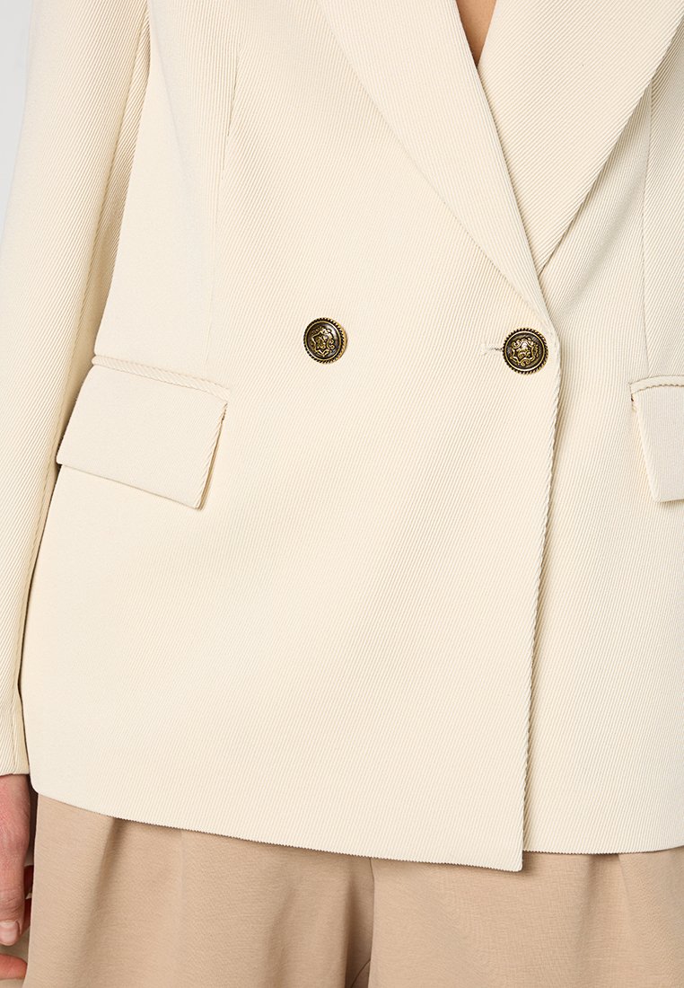 Blazer doppiopetto color crema in tessuto testurizzato, con revers a bavero, due tasche frontali e bottoni rotondi dorati.