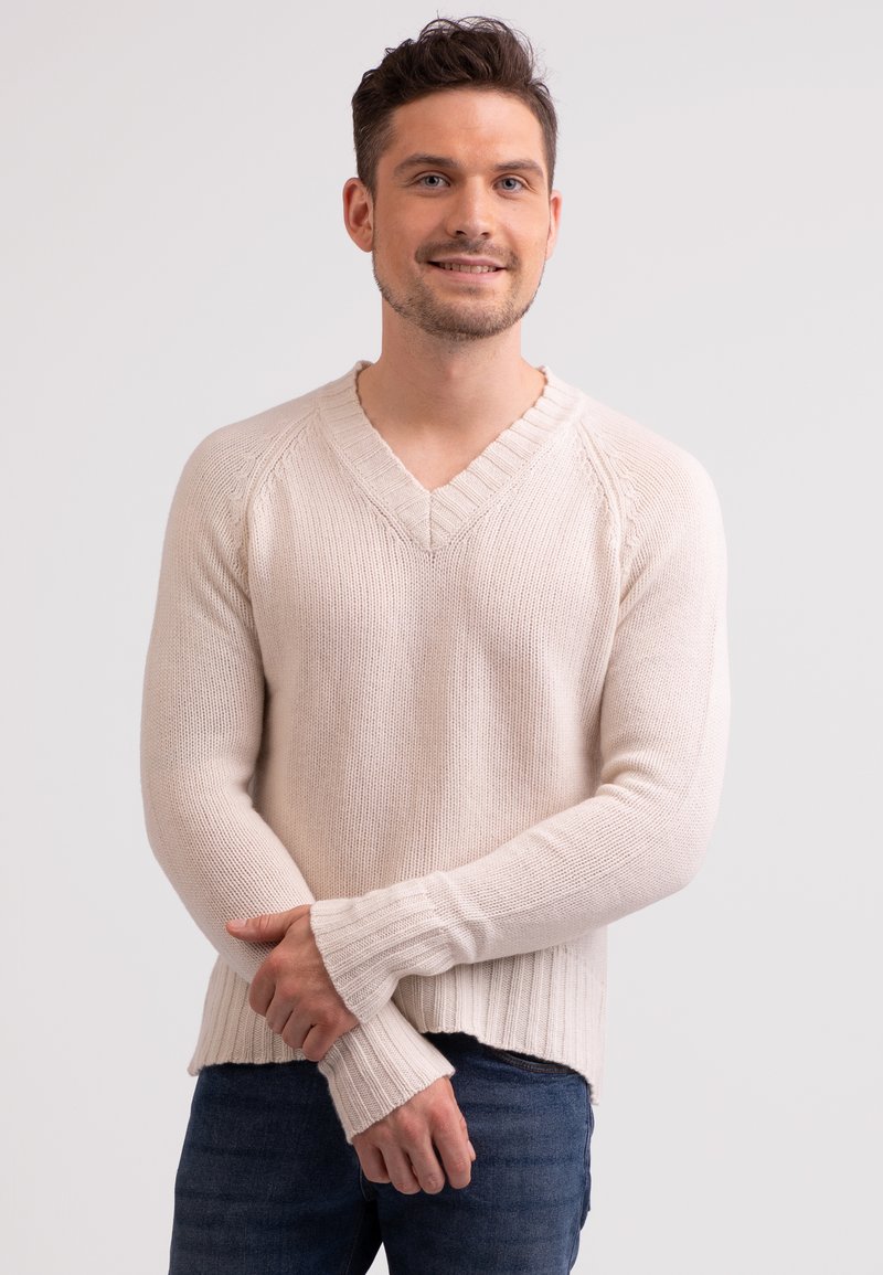 Hellbeige gestrickter Pullover mit V-Ausschnitt, gerippten Bündchen und einem etwas breiteren gerippten Saum. Die Textur wirkt weich und lässig.