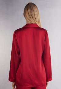 Röd satin långärmad skjorta med rak skärning, klassisk krage och slät, glänsande yta. Bakvy visar ärmslutar och kjol.