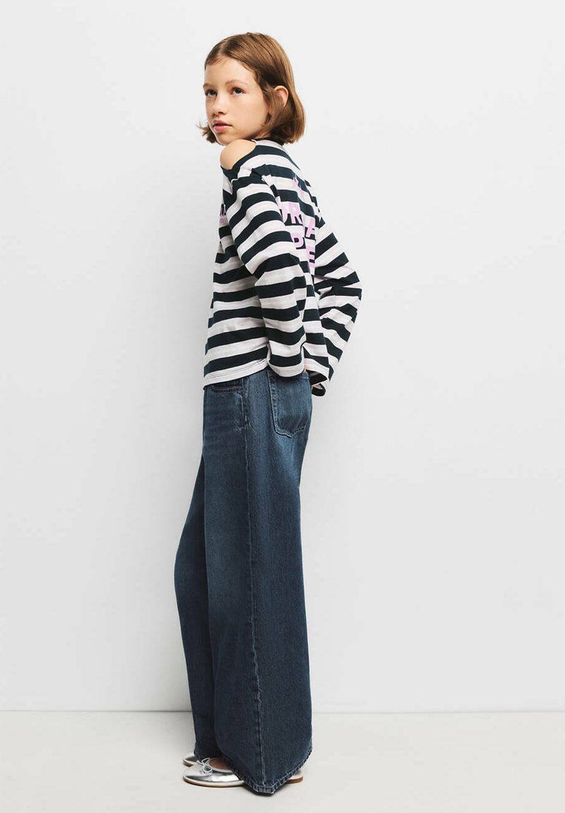 Jeune personne aux cheveux courts portant un top rayé noir et blanc à épaules dénudées, un jean large foncé et des chaussures argentées, debout de profil.