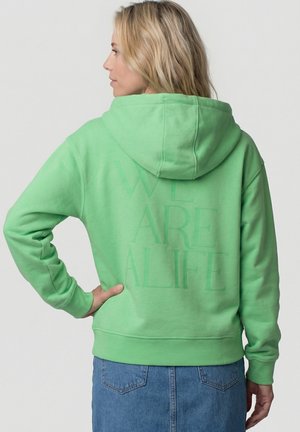 Blonde Frau im Profil, trägt einen hellgrünen Hoodie mit aufgesetzter Kapuze und dem Schriftzug "WE ARE ALIFE" auf dem Rücken, kombiniert mit einem blauen Jeansrock.