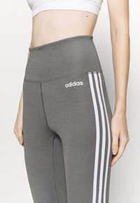 Leggings grigi alti in vita con strisce laterali bianche, realizzati in un tessuto morbido e testurizzato, con un logo Adidas ricamato sul davanti.