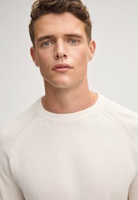 Weißer gestrickter Pullover mit strukturiertem Muster, geripptem Halsausschnitt und Raglanärmeln. Der Stoff scheint leicht und atmungsaktiv zu sein.
