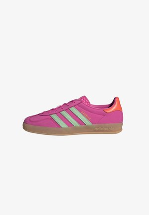 Vue de côté d'une chaussure de sport Adidas Gazelle rose avec des bandes vert clair, un languette orange au talon et une semelle en gomme.