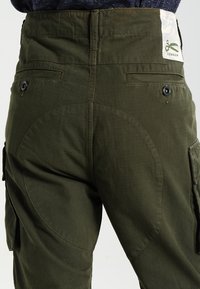 Olivegröna cargobyxor har bakfickor, böjda sömdetaljer och en märkesetikett. Tyget har en slät, strukturerad finish.