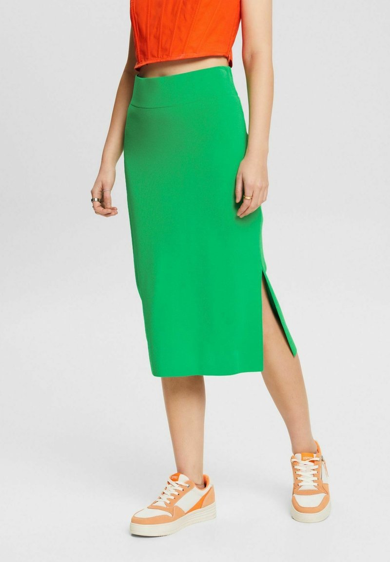Femme portant une jupe midi vert vif avec une fente latérale, un top court orange et des baskets orange et blanches, debout sur un fond gris uni.