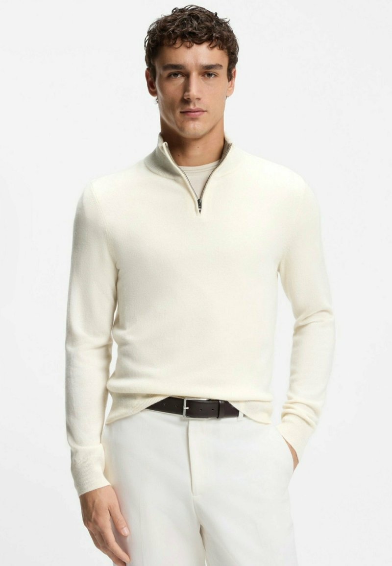 Heller, cremefarbener Pullover mit Viertel-Reißverschluss aus weichem Strickmaterial. Verfügt über lange Ärmel und eine glatte Textur, getragen mit dunklem Gürtel und hellem Hose.