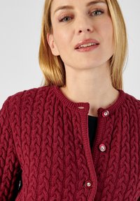 Gilet en tricot bourgogne avec un motif de câble texturé, encolure ronde et deux boutons rouges à l'avant. La douceur du fil est évidente dans le design.