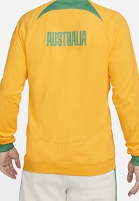 Gul atletisk jakke med grønne detaljer. "AUSTRALIEN" trykt på ryggen. Glat tekstur, lange ærmer og en afslappet pasform.