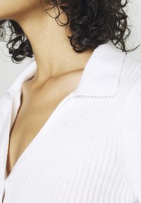 Vit ribbad stickad topp med platt krage och v-ringning, med texturerad tyg och subtila vertikala linjer genom hela.