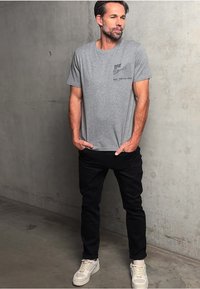 Muž v šedém grafickém tričku, černých kalhotách a bílých teniskách stojí s rukama v kapsách proti betonové zdi.