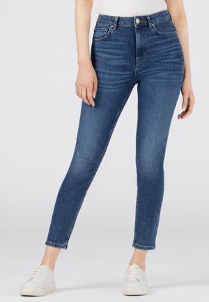 Femme portant un jean skinny bleu taille haute et des baskets blanches, debout, les pieds écartés sur un fond clair uni.