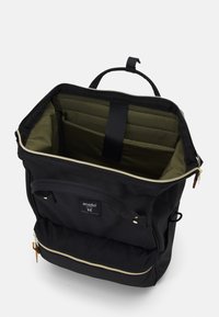 Sac à dos noir avec une fermeture éclair dorée et une poche avant. L'intérieur est doublé de vert et comprend une section rembourrée pour l'organisation.