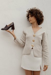 Zapato de tacón alto de cuero marrón con punta cuadrada y tacón grueso, sostenido por una persona que lleva una chaqueta beige y una falda blanca frente a una pared lisa.