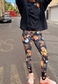 Sweat-shirt noir avec un graphique de soleil, associé à un legging à motifs floraux multicolores sur un pavement noir texturé, et des baskets rose pâle.