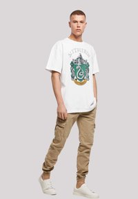 Hvit kortermet t-skjorte med et grønt Slytherin-våpendesign, kombinert med beige cargo-bukser og hvite joggesko. Enkelt bakgrunn.