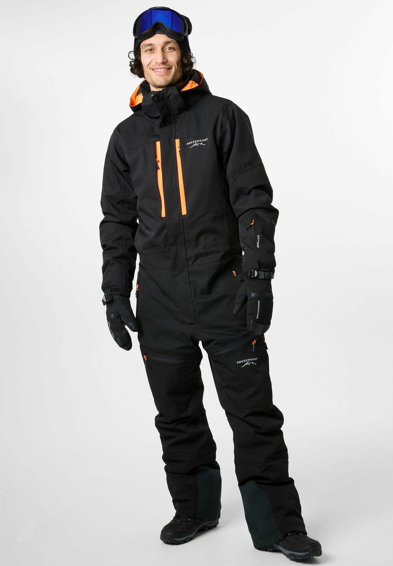 Zwarte sneeuwpak met oranje accenten, voorzien van ritszakken, een hoge kraag en handschoenen. Het model draagt een skibril en laarzen.