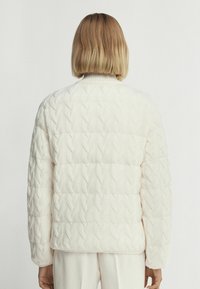 Vit quiltad jacka med kabelstickad textur, hög krage och avslappnad passform. Har puffade axlar och ribbade ärmslut.