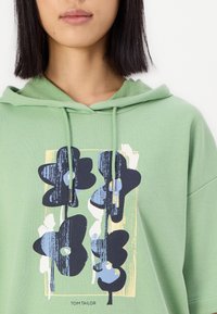 Groene hoodie met een trekkoord, versierd met een bloemenprint in marineblauw en lichtblauw. Zachte stof, oversized pasvorm en opvallend logo aan de onderkant.