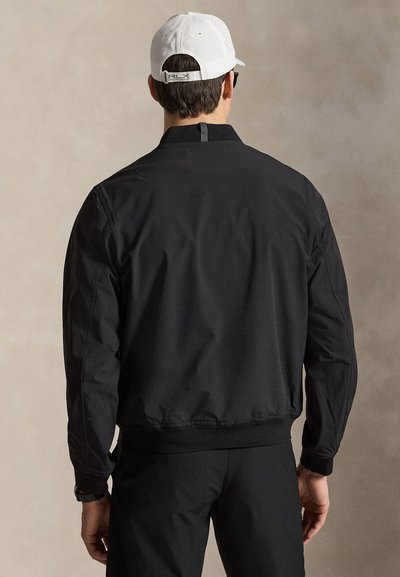 Muž v černé bundě a kalhotách s bílou baseballovou čepicí, stojící zády k jednobarevnému béžovému pozadí.