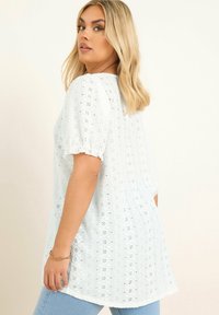 Blusa bianca in pizzo con maniche corte a sbuffo, superficie texturizzata, collo rotondo e vestibilità rilassata; abbinata a jeans di denim blu chiaro.