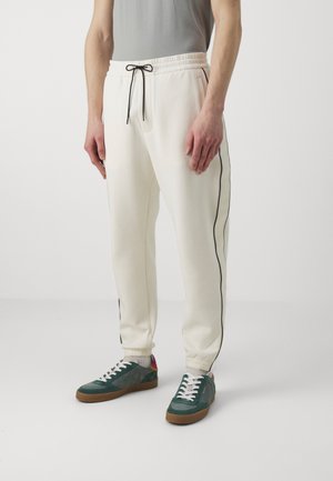 White sweatpants with black side piping, drawstring waist, and cuffed ankles, paired with green and gray sneakers featuring white laces.