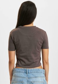 Brown, cropped t-shirt with short sleeves and a round neckline, made of soft material, displayed from the back, paired with light blue denim shorts.