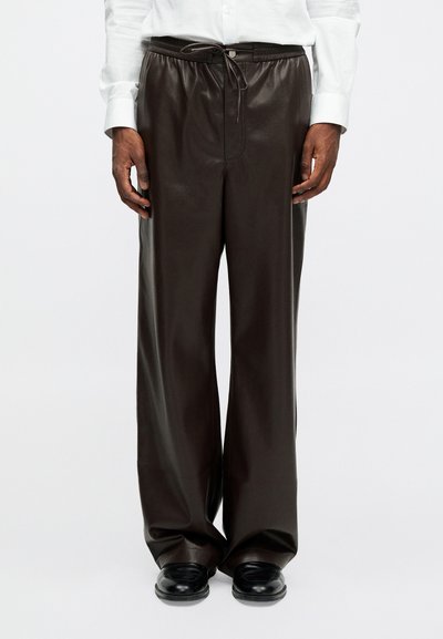 Moški nosi široke usnjene hlače temno rjave barve z vrvico v pasu in črne loafer čevlje, prikazan od pasu do nog pri belem ozadju.