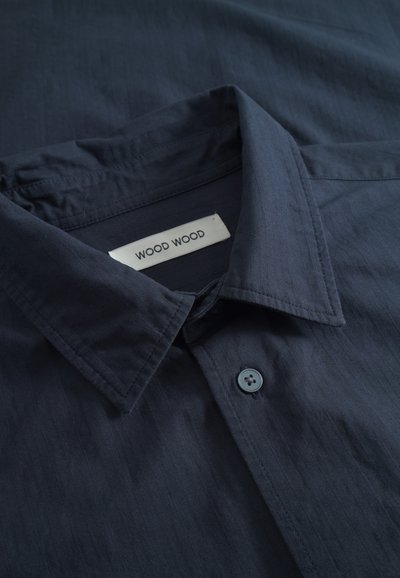 Camisa de botones azul marino con cuello puntiagudo, con una etiqueta de la marca "WOOD WOOD" cosida en el interior del escote.