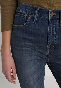 Dark blue denim jeans with a mid-rise waist, featuring a front pocket, copper button detail, and subtle texture.