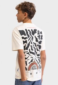 Beige bomulds t-shirt med et sort abstrakt bladdesign på ryggen samt teksten "Grow Into The Wild" og "Rebel" i rød.