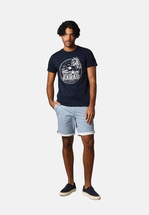 Marine t-shirt met "HAWAII" grafiek, lichtblauwe gestreepte shorts met opgerolde randen, en zwarte instap schoenen.