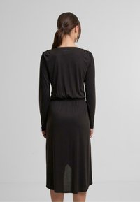 Schwarz langärmliges Kleid mit tailliertem Oberteil, elastischem Bund und fließendem Rock. Glatte Textur und dezente Raffung an den Schulternähten.