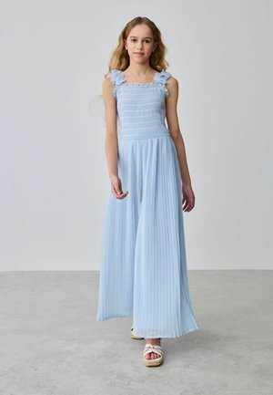 Dziewczyna o długich blond włosach, ubrana w bezrękawnikową, jasnoniebieską plisowaną suknię maxi i białe sandały, stojąca na szarej podłodze na tle jednolitego tła.