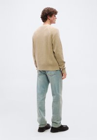 Jeune homme aux cheveux bouclés portant un pull en tricot beige, un jean bleu clair et des chaussures noires, debout devant un fond blanc uni.