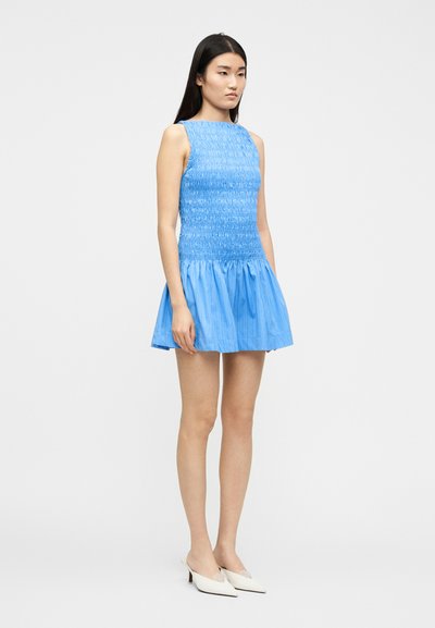 Γυναίκα όρθια, φορώντας κοντό μπλε φόρεμα χωρίς μανίκια με πιετά φούστα και άσπρα πέδιλα με αιχμηρή μύτη και λουράκι στο πίσω μέρος.