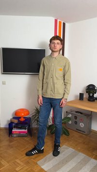 Kakhi kordbársony dzseki, két elülső zseb, gombos záródás és címke; kék farmernadrággal és fekete sportcipővel párosítva. Modern beltéri környezet.