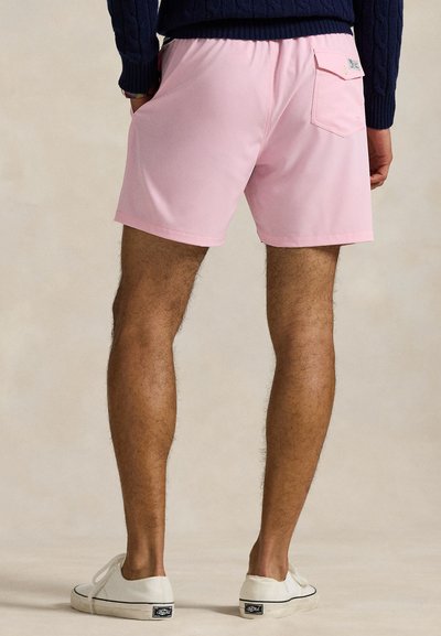 Moški v svetloružičastih kratkih hlačah z zadnjim žepom, temno modrem puloverju in belih supergah stoji ob običajni podlagi.