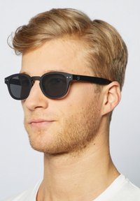 Czarne okulary przeciwsłoneczne z okrągłymi oprawkami, matowym wykończeniem, szarymi soczewkami i małymi srebrnymi akcentami na zausznikach. Noszone przez młodego mężczyznę o jasnych włosach.