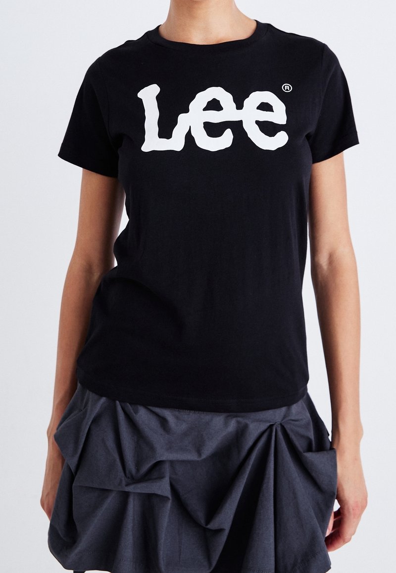 Personne portant un t-shirt noir avec un logo blanc "Lee" et une jupe gris foncé avec des détails de tissu plissé, debout devant un fond uni.