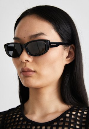 Czarne okulary przeciwsłoneczne w kształcie kocich oczu z ciemnymi soczewkami, błyszcząca plastikowa oprawka i złote akcenty z logo na skroni. Minimalistyczny design i lekka tekstura.