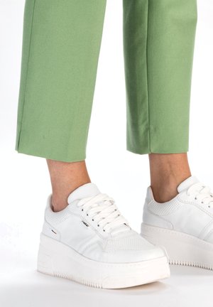 Baskets blanches avec une tige en cuir texturé et des accents perforés, dotées d'une épaisse semelle en caoutchouc blanche et de lacets blancs. Associées à un pantalon vert.