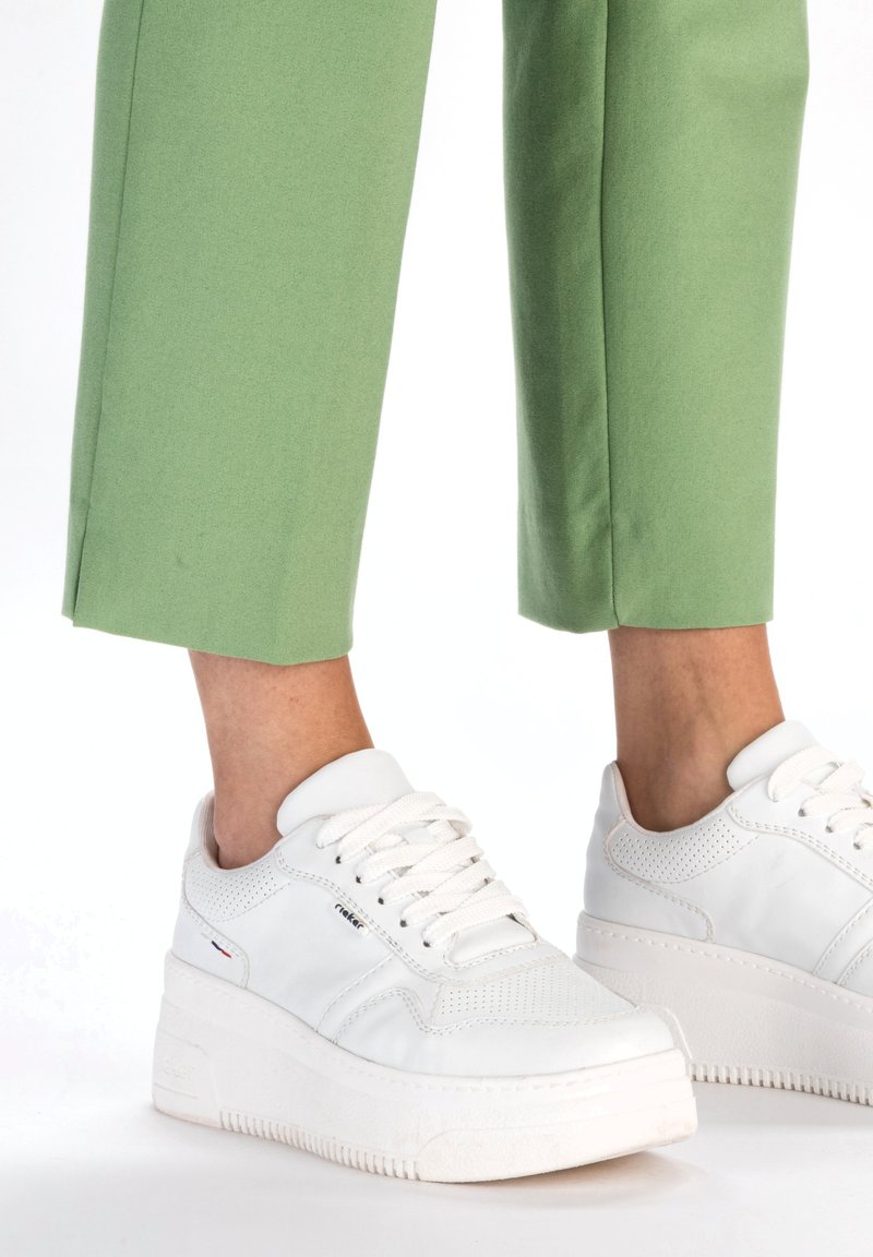Sneakers bianchi con tomaia in pelle testurizzata e accenti perforati, dotati di una suola in gomma spessa bianca e lacci bianchi. Abbinati a pantaloni verdi.