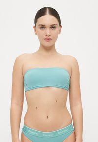 Τουρκουάζ μπικίνι τοπ σε μπαντό, με αντίστοιχα χαμηλοκάβαλα σλιπ και λευκό λογότυπο Calvin Klein στην ελαστική ζώνη. Απαλή ύφανση, μινιμαλιστικός σχεδιασμός.