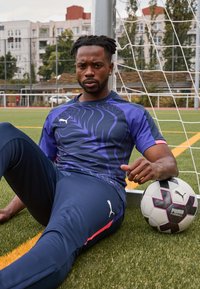 Moški v modri športni obleki sedi na trati, se naslanja na gol za nogomet in drži nogometno žogo Puma.