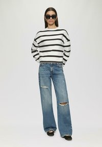 Crop top a maniche lunghe bianco e nero a righe abbinato a jeans larghi strappati di un leggero blu e scarpe nere. Caratterizzato da occhiali da sole neri oversize.