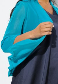 Kevyt, läpikuultava turkoosi huivi, jossa on leveät hihat, on drapattu tumman laivastonsinisen mekon päälle, korostaen sen sileää rakennetta ja virtaavaa muotoilua.