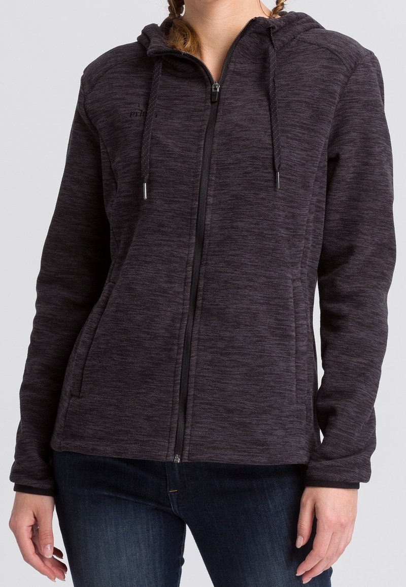 Femme portant un sweat à capuche zippé gris foncé avec une capuche et des cordons, associé à un jean bleu foncé, se tenant devant un fond neutre.