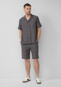 Korte mouw, button-up shirt in donkergrijze stof met een zak; gecombineerd met bijpassende shorts. Gdraagd met witte sneakers.
