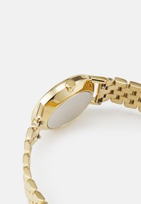 Zlatý kovový hodinky se hladkým zaobleným pouzdrem, malou korunou a náramkem z článků s střídavým oválným tvarem a leštěným povrchem.